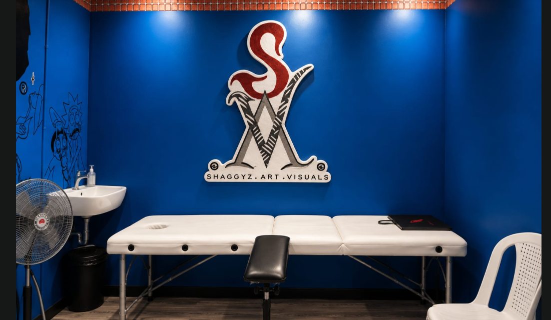 The Shaggyz Art Visuals sign mounted on the studio's blue wall above the tattoo bed
